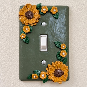 Op de afbeelding: Een rechthoekige olijfgroene lichtschakelaarplaat met een witte tuimelschakelaar in het midden en twee zilveren schroeven. De plaat is versierd met driedimensionale gele zonnebloemen, kleinere gele bloemen met witte harten en groene bladeren.