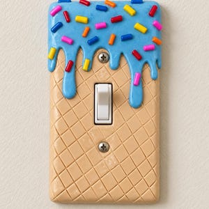 Op de afbeelding: Een lichtschakelaarplaat ontworpen als een ijshoorntje. Het onderste gedeelte heeft een beige wafelpatroon, terwijl de bovenkant is bedekt met blauw glazuur en kleurrijke hagelslag. Een witte tuimelschakelaar is in het midden geplaatst.