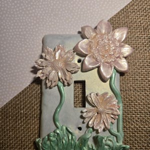 Op de afbeelding: Decoratieve lichtschakelaar cover met roze bloemen en groene stelen en bladeren. De cover is lichtblauw. De achtergrond is lichtroze en bruin. De schakelaarplaat meet ongeveer 12 cm hoog.