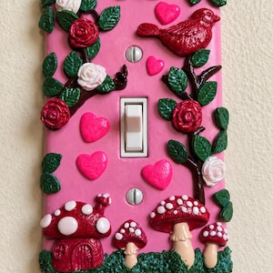 Op de afbeelding: Een rechthoekige roze lichtschakelaarafdekking met driedimensionale decoraties, waaronder rode en witte rozen, groene bladeren, rode harten, een rode vogel en verschillende rood-witte stippenpaddenstoelen aan de onderkant met een witte schakelaar.