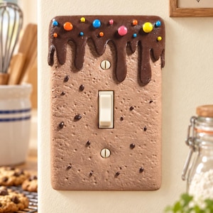 Op de afbeelding: Een rechthoekige lichtschakelaar-afdekplaat ontworpen als een koekje met chocoladeglazuur en kleurrijke suikerkorrels. Het getextureerde oppervlak heeft een lichtbruine koekjesbasis met donkerbruine druppels aan de bovenkant en een tuimelschakelaar.