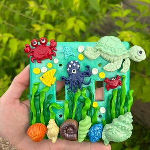 May include: A decorative light switch cover featuring an underwater scene. The cover is teal with sculpted sea creatures, including a red crab, a yellow fish, a blue octopus, and a green turtle. Seashells and seaweed are also present.