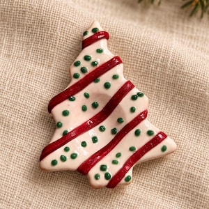 Könnte beinhalten: Ein Weihnachtsbaumschmuck aus Keramik mit einer glänzenden cremefarbenen Glasur, verziert mit diagonalen dunkelroten Streifen und kleinen, erhabenen grünen Punkten, die über die Oberfläche verteilt sind.