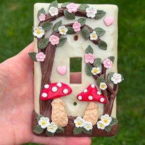 May include: A rectangular light switch plate with a cream-colored base featuring a raised, three-dimensional design of two red-capped mushrooms with white spots, brown tree branches, green leaves, small white and pink flowers, and two pink hearts.