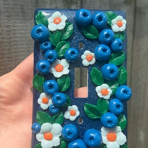 Op de afbeelding: Een rechthoekige donkerblauwe lichtschakelaarplaat met een driedimensionaal decoratief patroon van gebeeldhouwde bosbessen, groene bladeren en kleine witte bloemen met oranje harten verspreid over het glinsterende oppervlak.