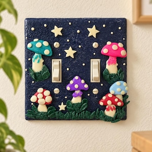 Op de afbeelding: Een donkerblauwe dubbele lichtschakelaarplaat met een getextureerd oppervlak, voorzien van verhoogde, kleurrijke paddenstoelfiguren in rood, roze, paars en blauw, geaccentueerd door kleine witte sterren en groen blad rond de voet.