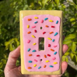Op de afbeelding: Een decoratieve lichtschakelaarcover die lijkt op een glazuurgebakje. De cover heeft een gele basis met een roze glazuurlaag en kleurrijke hagelslag. De schakelaaropening is rechthoekig.