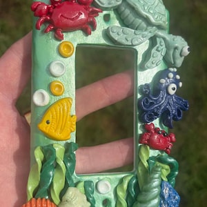 Op de afbeelding: Een rechthoekige lichtschakelaar-afdekplaat met een metallic groene afwerking, versierd met driedimensionale zeedieren, waaronder een rode krab, een lichtgroene zeeschildpad, een donkerblauwe octopus, een gele vis, schelpen en groen zeewier.