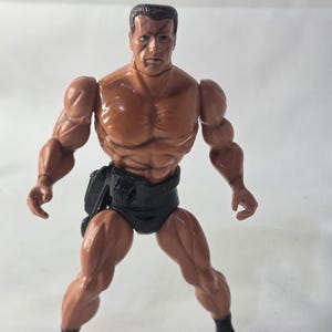 May include: A muscular action figure with a tan complexion, short dark hair, and a visible scar over the left eye. The figure wears black trunks and black boots, standing in a wide stance with arms slightly bent at the sides and defined muscles.