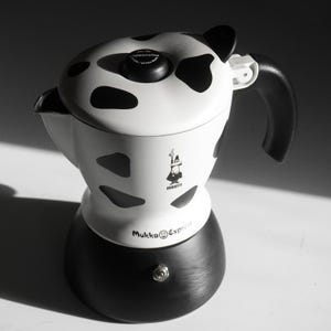 Puede incluir: Una cafetera de estufa Bialetti Mukka Express con una cámara superior blanca con manchas negras de vaca, una base negra y un asa negra. La tapa incluye un botón central con la etiqueta cappuccino y el cuerpo muestra el logotipo de Bialetti.