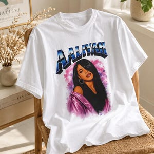 May include: A white short-sleeved t-shirt featuring a graphic print of the singer Aaliyah. The design includes the name AALIYAH in a metallic blue font above a portrait of the artist wearing a purple jacket, set against a pink and purple nebula background.