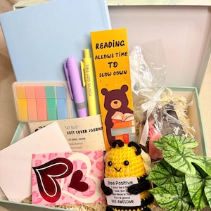 May include: A gift box containing reading-related items. It features a light blue journal, a bookmark with a bear reading, pens, sticky notes, a crochet bee holding a sign saying "Bee Positive", and a card with heart designs.