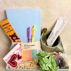 May include: A gift box containing a sky blue journal, pens, sticky notes, a bookmark, a mug with a caramel latte sachet, and a decorative spoon. The bookmark reads "Reading Allows Time to Slow Down". The journal is labelled "Soft Cover Journal".