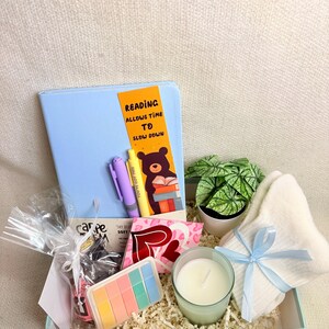 May include: A gift box containing a light blue journal, pens, a small potted plant, a candle, socks, and other small gifts. The journal has a bookmark with the text "Reading Allows Time to Slow Down". The box is filled with shredded paper.