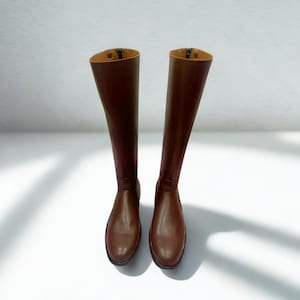 以下が含まれることがあります： 滑らかな仕上げの膝丈のブラウンレザーブーツ一足。丸みを帯びたトゥとタンカラーの裏地が特徴です。ブーツのシャフトには縦の縫い目があり、右上隅には「HIDE CRAFT LEATHER HOUSE STORE」と書かれたロゴが配置されています。
