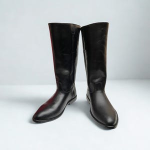 Könnte beinhalten: Ein Paar kniehohe schwarze Stiefel mit glatter Oberfläche, abgerundeten Zehen und einem niedrigen Absatz. Die Stiefel haben ein klares, strukturiertes Design mit sichtbaren Nähten an den Seiten und einem Logo oben rechts mit der Aufschrift HIDE CRAFT.