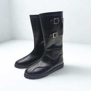 May include: A pair of black mid-calf boots featuring two horizontal straps with silver-toned metal buckles on the outer side. The boots have a smooth finish, a rounded toe, and a flat sole. The text HIDE CRAFT is visible in the top right corner.