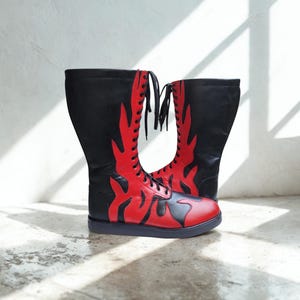Puede incluir: Un par de botas de media caña con un patrón de llamas en negro y rojo. Las botas tienen cordones negros, punta redondeada y una suela plana negra. La parte superior es principalmente negra con superposiciones de llamas rojas.