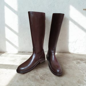 May include: A pair of knee-high, brown leather boots. The boots have a smooth, polished finish and a classic design. They are standing against a white wall, with shadows from natural light. The boots are a rich, dark brown colour.