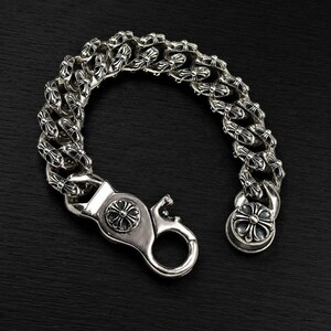 May include: A silver bracelet with a curb chain design. Each link features a cross-like pattern. The bracelet has a large clasp with a decorative circular element and a smaller clasp on the other end. The bracelet is displayed against a dark background.