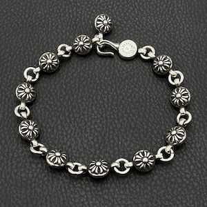 May include: Silver bracelet with daisy-shaped floral elements. The bracelet is linked with small, curved connectors and fastens with a hook clasp. The bracelet is made of a shiny, metallic material.