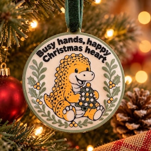 May include: A round, cream-colored Christmas ornament featuring a yellow pangolin holding a small green tree. The text "Busy hands, happy Christmas heart" is printed in black above the pangolin, framed by green holly leaves and berries.