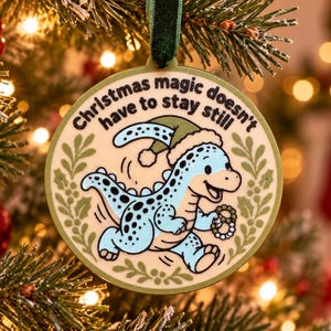 May include: A round, cream-colored ornament featuring a light blue dinosaur wearing a green Santa hat and holding a wreath. The text "Christmas magic doesn't have to stay still" is printed in black above the dinosaur, framed by green holly leaves.