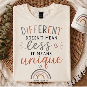May include: A cream-colored t-shirt featuring the text "DIFFERENT DOESN'T MEAN less IT MEANS unique" in various colors and fonts. The design includes small heart icons and a multi-colored rainbow graphic at the bottom with the text "CHAOS & CALM CO."