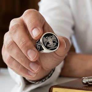 May include: A silver signet ring featuring a black circular inlay with a detailed silver double-headed eagle emblem and the text "SCOTTISH RITE" engraved below the eagle. The ring has a polished metallic band and a flat, circular face design.