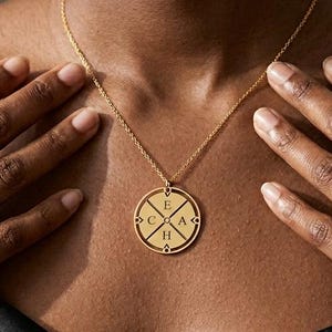 May include: A gold-colored pendant necklace with a circular design featuring the letters E, C, A, and H. The necklace hangs on a delicate chain. The person is wearing a black satin top and gold rings.