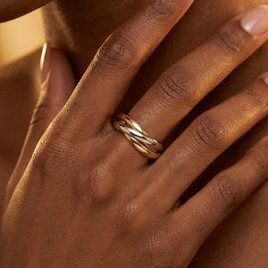 Può includere: Un anello dorato e argentato con un design intrecciato. L'anello è indossato su un dito, con la mano e parte del braccio visibili. L'incarnato è medio e lo sfondo è sfocato.