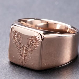 May include: A rose gold-toned signet ring featuring a polished, smooth surface and a detailed, engraved design of symmetrical wings on the flat, square-shaped face. The metal has a warm, metallic luster with visible reflections on the curved band.