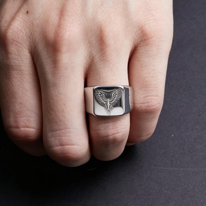 May include: A silver-toned signet ring featuring a flat, square face engraved with a detailed illustration of spread wings. The metal has a polished, reflective finish, and the wings are depicted with fine, dark lines against the metallic surface.