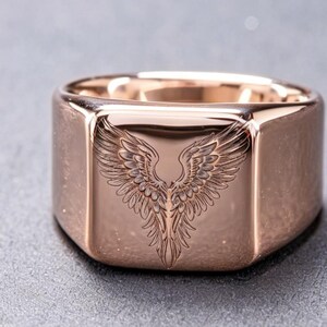 May include: A rose gold-toned signet ring featuring a polished, smooth surface with a deeply engraved, detailed illustration of a pair of symmetrical bird wings centered on the flat, square-shaped face of the band.