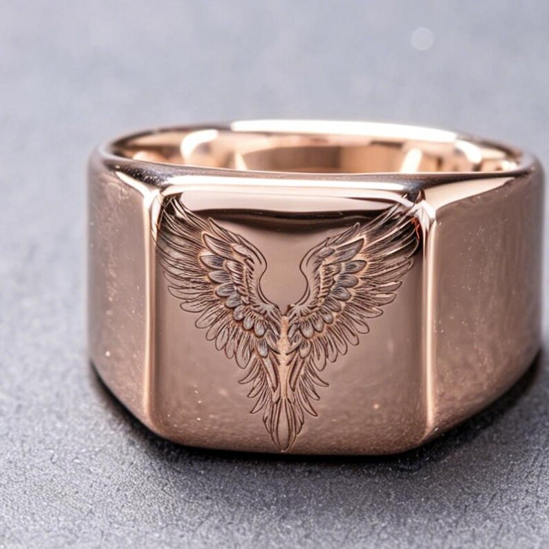 May include: A rose gold-toned signet ring featuring a polished, smooth surface with a deeply engraved, detailed illustration of a pair of symmetrical bird wings centered on the flat, square-shaped face of the band.