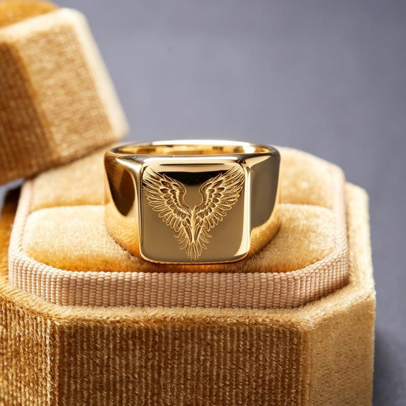 May include: A polished gold signet ring featuring a square face with a detailed, engraved design of symmetrical wings spreading upward. The ring has a smooth, reflective surface and is centered within a textured, golden-colored velvet box.