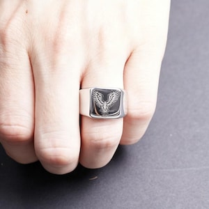 May include: A silver-toned signet ring featuring a flat, rectangular face with a black inlay. The inlay is engraved with a detailed white graphic of a bird with wings spread wide, positioned symmetrically in the center of the ring's face.