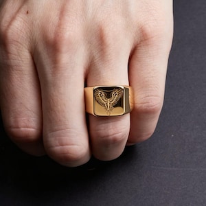 May include: A gold-toned signet ring featuring a flat, square face with an engraved design of a bird with outstretched wings. The ring has a polished, reflective surface and a wide, solid band that wraps around the finger.