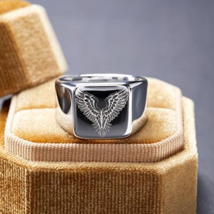 May include: A polished silver-tone signet ring featuring a flat, square face with a detailed, laser-engraved design of symmetrical bird wings. The metal surface is highly reflective, showcasing a smooth, rounded band and a clean, metallic finish.