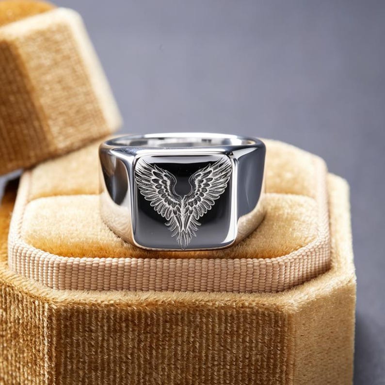 May include: A polished silver-tone signet ring featuring a flat, square face with a detailed, laser-engraved design of symmetrical bird wings. The metal surface is highly reflective, showcasing a smooth, rounded band and a clean, metallic finish.