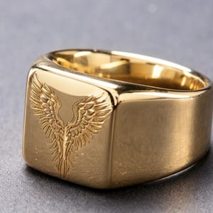 May include: A gold-toned signet ring featuring a polished, smooth surface and a square face. The front of the ring is engraved with a detailed, symmetrical design of two spread wings meeting in the center, creating a feathered pattern.