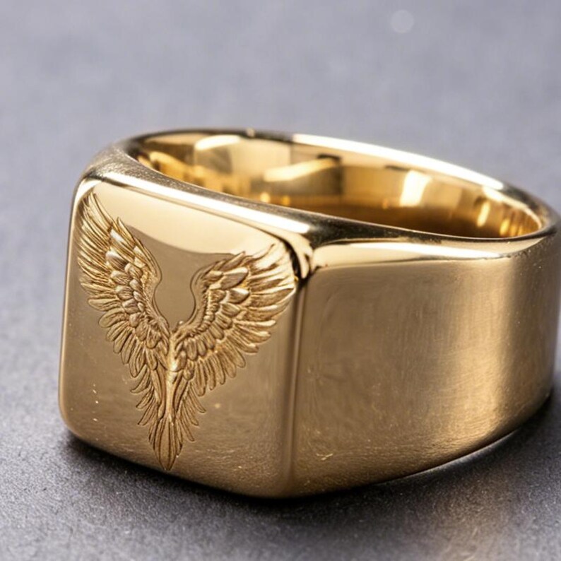 May include: A gold-toned signet ring featuring a polished, smooth surface and a square face. The front of the ring is engraved with a detailed, symmetrical design of two spread wings meeting in the center, creating a feathered pattern.