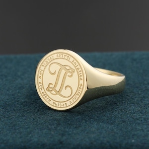 May include: A gold signet ring featuring a circular face with a polished finish. The center of the ring displays an engraved cursive letter L, surrounded by a border containing a series of small, repeating geometric symbols etched into the metal.