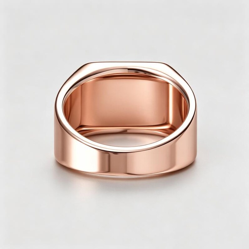 May include: A rose gold signet ring with a smooth, polished finish and a wide, flat top surface. The band is solid and features a rounded interior profile, showcasing a metallic luster and a clean, minimalist design with no engravings or stones.