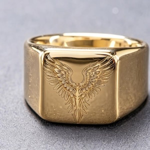 May include: A gold-toned signet ring featuring a flat, square face engraved with a detailed pair of symmetrical wings. The polished metal surface reflects light, highlighting the intricate feather textures etched into the center of the ring.