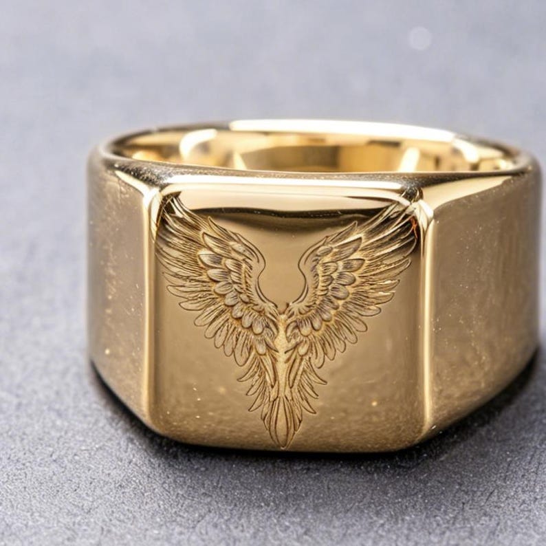 May include: A gold-toned signet ring featuring a flat, square face engraved with a detailed pair of symmetrical wings. The polished metal surface reflects light, highlighting the intricate feather textures etched into the center of the ring.