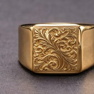 Pode incluir: Um anel de sinete dourado com uma face quadrada. A face do anel apresenta um padrão floral ornamentado e rodopiante. O anel tem um acabamento polido e uma banda grossa.