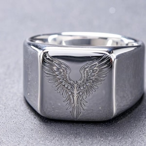May include: A polished silver-tone signet ring featuring a square face with a detailed, engraved design of symmetrical bird wings spreading upward. The metal surface is smooth and reflective, showcasing the intricate feather textures of the wings.
