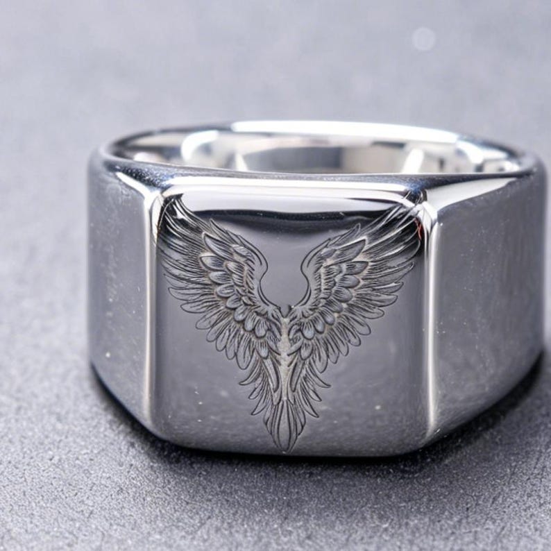 May include: A polished silver-tone signet ring featuring a square face with a detailed, engraved design of symmetrical bird wings spreading upward. The metal surface is smooth and reflective, showcasing the intricate feather textures of the wings.