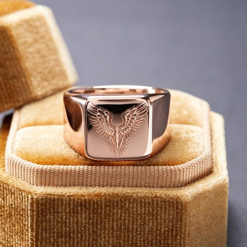 May include: A rose gold signet ring featuring a polished finish and a detailed, engraved design of symmetrical wings on the flat square face. The ring has a smooth, solid band and a reflective surface that catches the light.